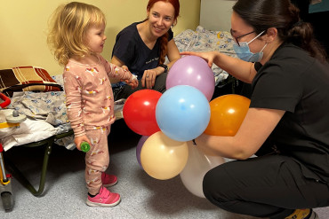 Pracownik Szpitala kucając przed małą dziewczynką daje jej balona, obok dziewczynki siedzi jej mama na łóżku szpitalnym