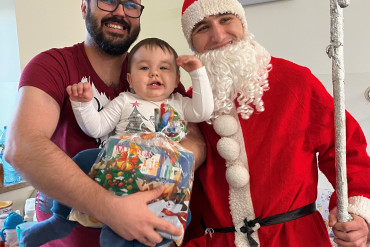 Na zdjęciu stoi tata trzymający małe dziecko na rękach, obok święty Mikołaj