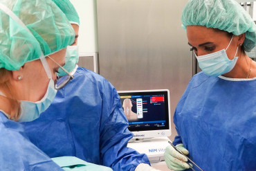 Lekarze na sali operacyjnej ubrani w odzież medyczną, za nimi urządzenie do neuromonitoringu.
